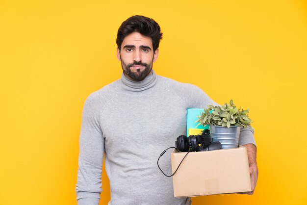 Jovem bonito com barba, fazendo um movimento ao pegar uma caixa cheia de coisas ao longo da parede isolada com expressão triste