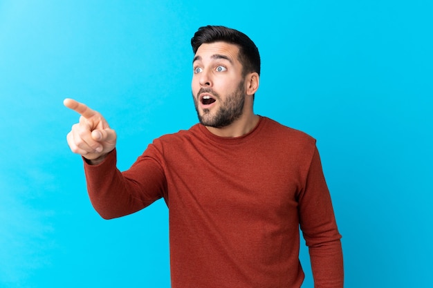 Jovem bonito com barba isolada, fundo azul apontando para longe