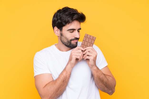 Jovem bonito com barba isolado parede amarela tomando uma tablete de chocolate e feliz