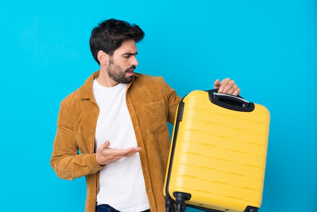 Jovem bonito com barba isolado parede azul em férias com mala de viagem