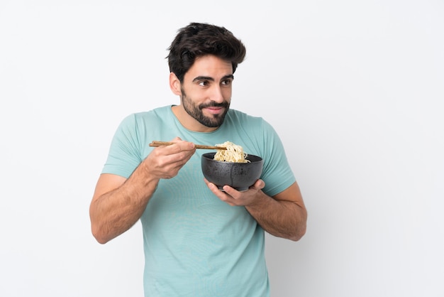 Jovem bonito com barba isolado parede branca segurando uma tigela de macarrão com pauzinhos