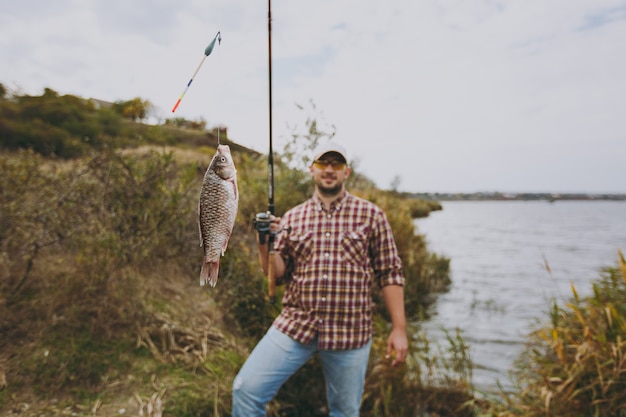 Jovem bonito com barba por fazer em camisa quadriculada, boné e óculos escuros segura uma vara de pescar com peixes capturados na margem do lago perto de arbustos e juncos. estilo de vida, recreação, conceito de lazer de pescador