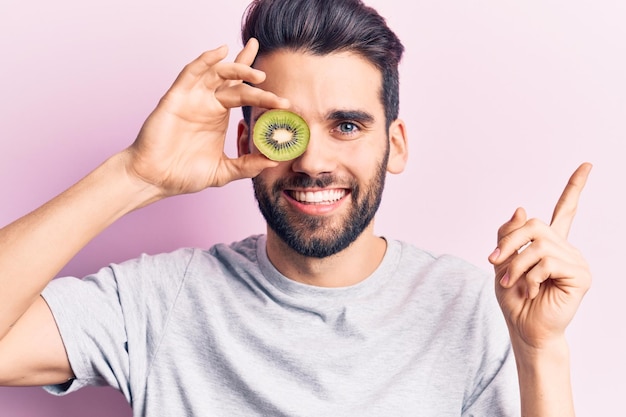 Jovem bonito com barba segurando kiwi sobre o olho sorrindo feliz apontando com a mão e o dedo para o lado