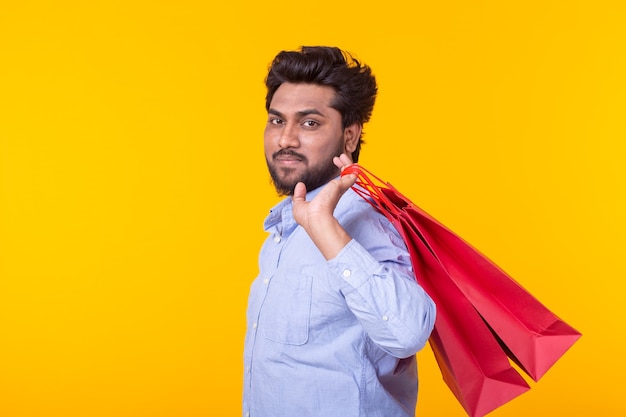 Jovem bonito com barba segurando sacolas vermelhas, posando em uma parede amarela