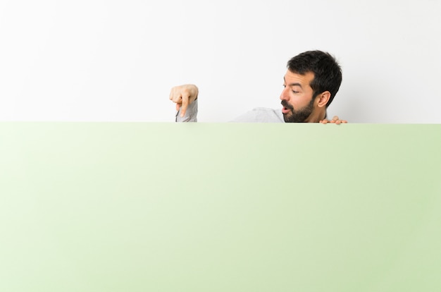 Jovem bonito com barba segurando um cartaz vazio verde grande e apontando-o