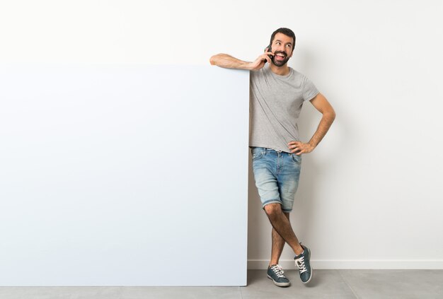 Jovem bonito com barba segurando um grande cartaz vazio azul, mantendo uma conversa com o telefone móvel