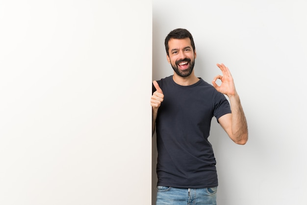 Jovem bonito com barba segurando um grande cartaz vazio mostrando sinal okey e polegar para cima gesto