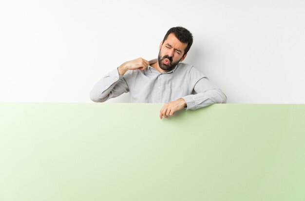 Jovem bonito com barba segurando um grande cartaz vazio verde com expressão cansada e doente