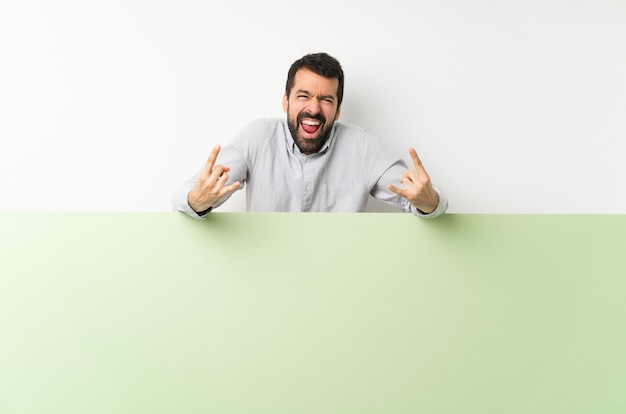 Jovem bonito com barba segurando um grande cartaz vazio verde fazendo gesto de pedra