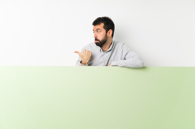 Jovem bonito com barba segurando um grande cartaz vazio verde infeliz e apontando para o lado