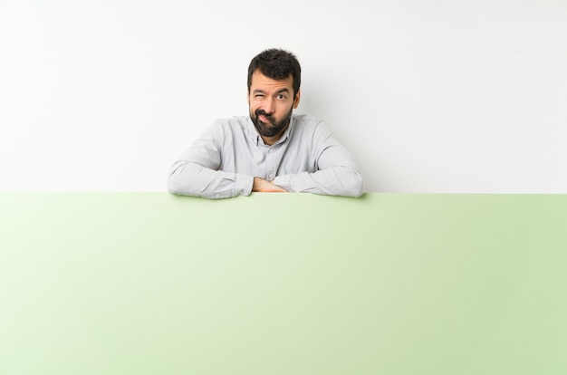 Jovem bonito com barba segurando um grande cartaz vazio verde sentindo chateado