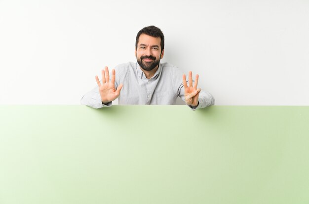 Jovem bonito com barba segurando um grande cartaz verde vazio, contando oito com os dedos