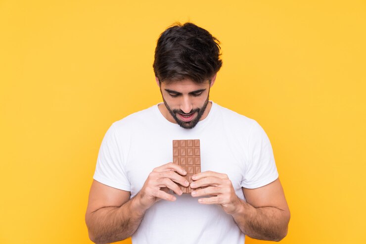 Foto jovem bonito com barba sobre parede amarela isolada, comendo uma tablete de chocolate