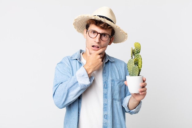 jovem bonito com boca e olhos bem abertos e mão no queixo. agricultor segurando um cacto decorativo