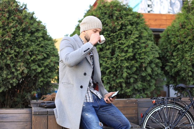 Jovem bonito com casaco cinza e chapéu, sentado num banco relaxado, bebendo café e pensando perto de sua bicicleta.
