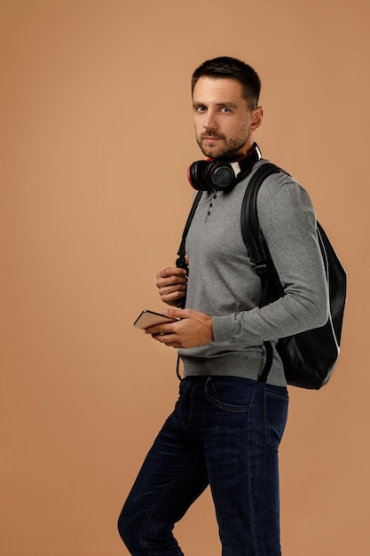 Foto jovem bonito com fones de ouvido e mochila usando smartphone isolado em fundo bege