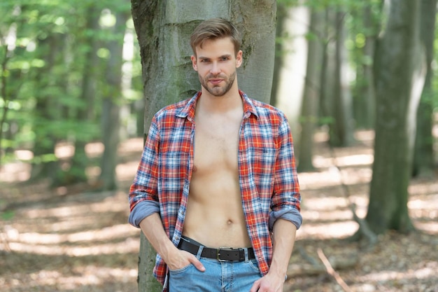 Jovem bonito com olhar lumbersexual em camisa desabotoada de lenhador em pé na árvore