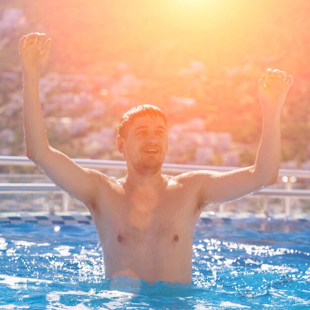 Jovem bonito com os braços erguidos à luz do sol da piscina em tons