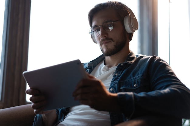 Jovem bonito com roupas casuais, usando um tablet digital e fones de ouvido e sorrindo enquanto está sentado na cadeira.