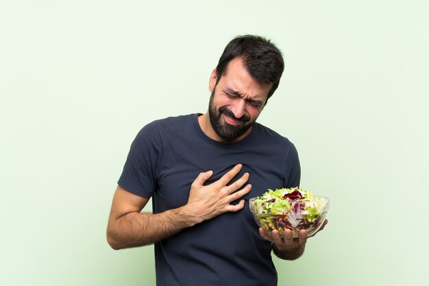 Jovem bonito com salada ao longo da parede verde isolada, com uma dor no coração