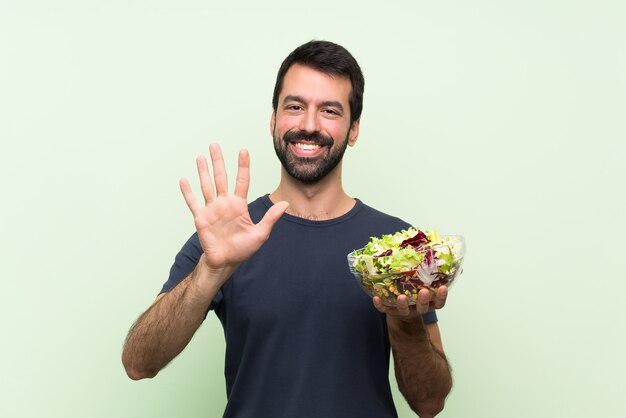 Jovem bonito com salada ao longo da parede verde isolada, contando cinco com os dedos