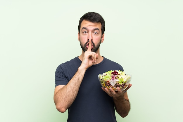 Jovem bonito com salada ao longo da parede verde isolada, mostrando um sinal de gesto de silêncio colocando o dedo na boca