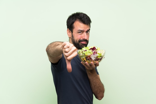 Jovem bonito com salada ao longo da parede verde, mostrando o polegar para baixo com expressão negativa