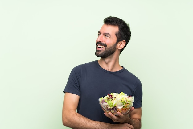 Jovem bonito com salada sobre parede verde isolada feliz e sorridente