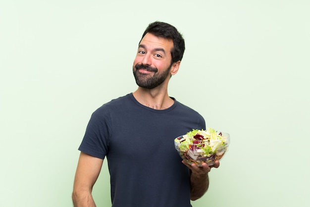 Jovem bonito com salada sobre parede verde isolada rindo