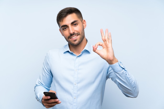 Jovem bonito com seu celular isolado parede azul mostrando sinal okey com os dedos