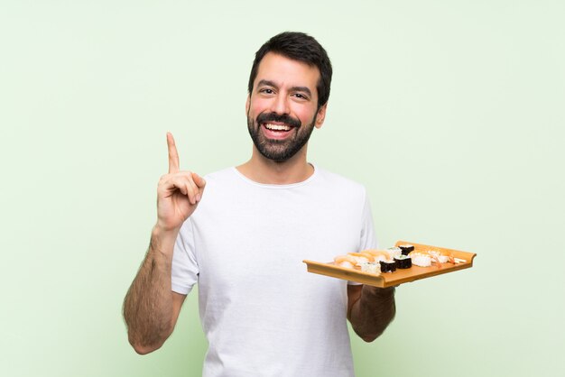 Jovem bonito com sushi sobre parede verde isolada, apontando para cima uma ótima idéia