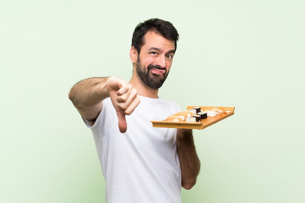 Jovem bonito com sushi sobre parede verde isolada, mostrando o polegar para baixo com expressão negativa