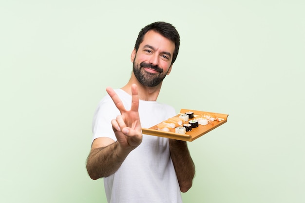 Foto jovem bonito com sushi sobre parede verde isolada, sorrindo e mostrando sinal de vitória