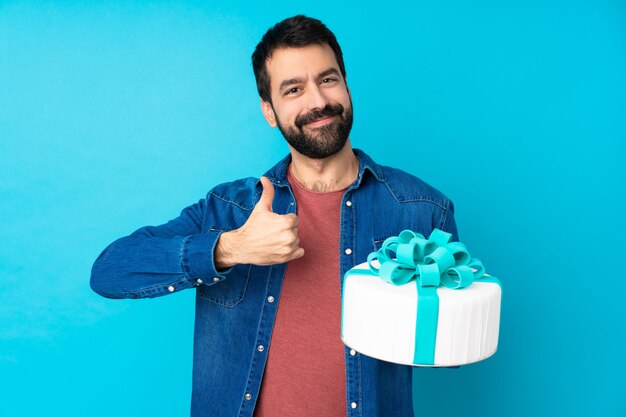 Jovem bonito com um bolo grande sobre parede azul isolada, dando um polegar para cima gesto