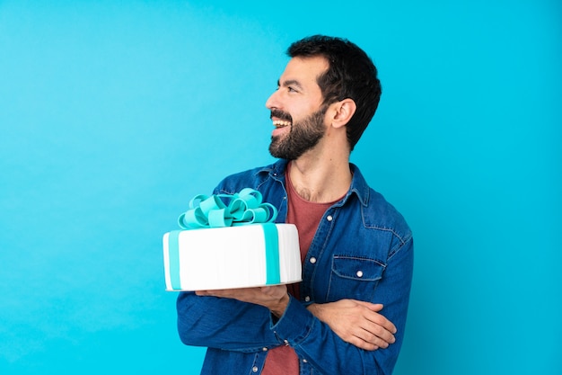 Jovem bonito com um bolo grande sobre parede azul isolada feliz e sorridente