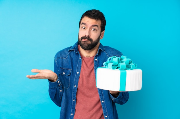 Jovem bonito com um bolo grande sobre parede azul isolada, tendo dúvidas ao levantar as mãos