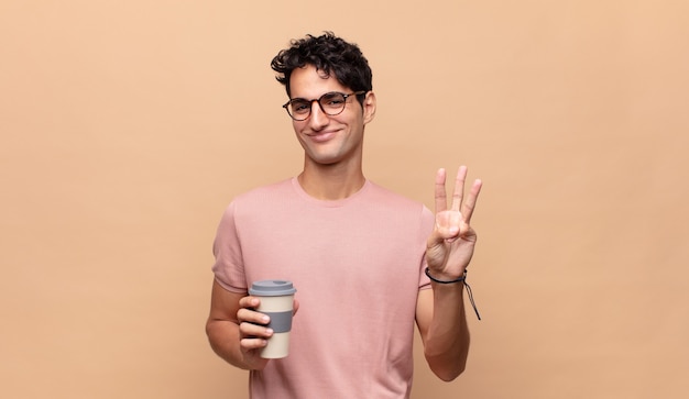 Jovem bonito com um café sorrindo e parecendo amigável, mostrando o número dois ou o segundo com a mão para a frente, em contagem regressiva