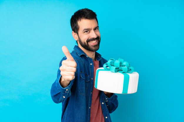 Jovem bonito com um grande bolo isolado parede azul com polegares para cima porque algo bom aconteceu