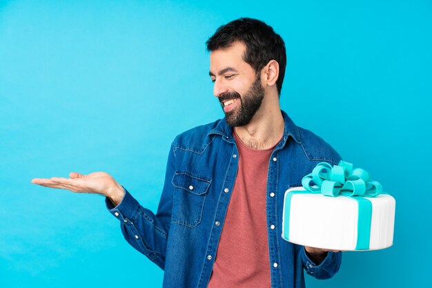 Jovem bonito com um grande bolo na parede azul, segurando o espaço em branco com as duas mãos