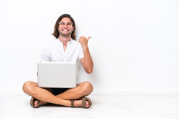 Jovem bonito com um laptop sentado no chão, isolado no fundo branco, apontando para o lado para apresentar um produto