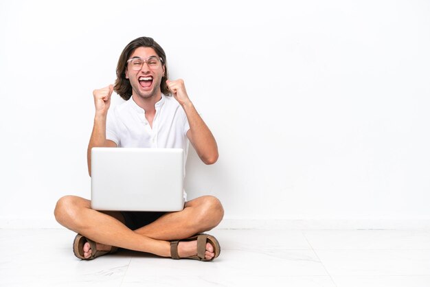 Jovem bonito com um laptop sentado no chão isolado no fundo branco comemorando uma vitória na posição de vencedor