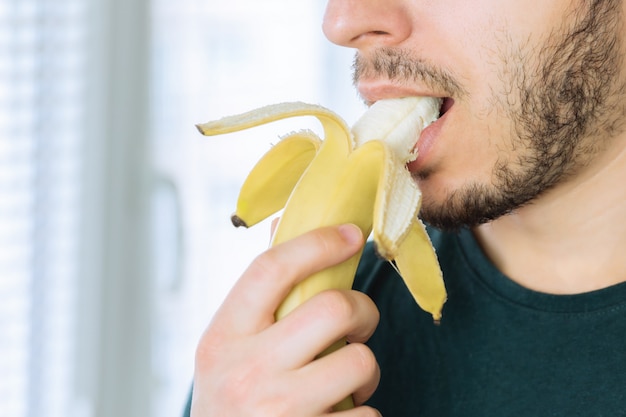 Jovem bonito com uma barba, mordendo uma banana em pé na cozinha.