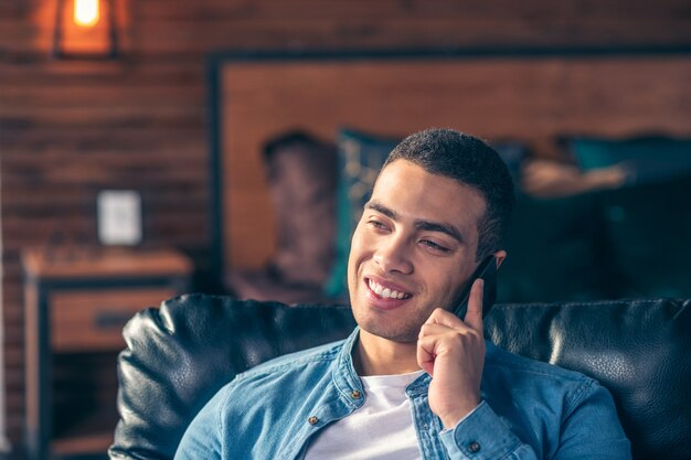 Jovem bonito com uma jaqueta jeans, falando em um smartphone e sorrindo.