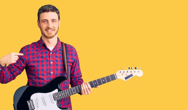 Jovem bonito com urso tocando guitarra elétrica apontando o dedo para si mesmo sorrindo feliz e orgulhoso