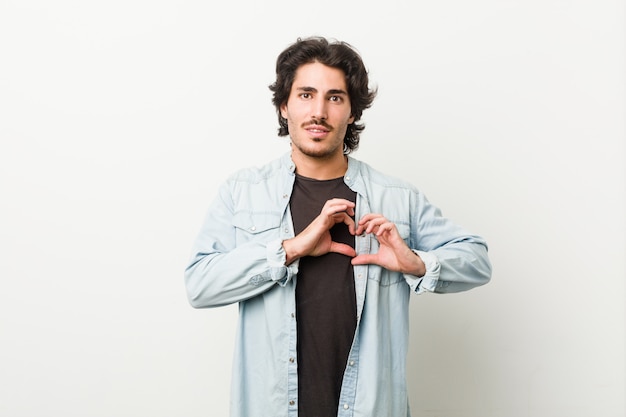 Jovem bonito contra um branco sorrindo e mostrando uma forma de coração com as mãos.