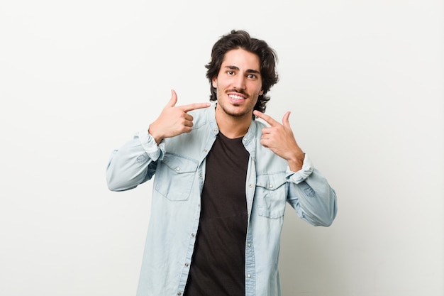 Jovem bonito contra uma parede branca sorri, apontando os dedos na boca.