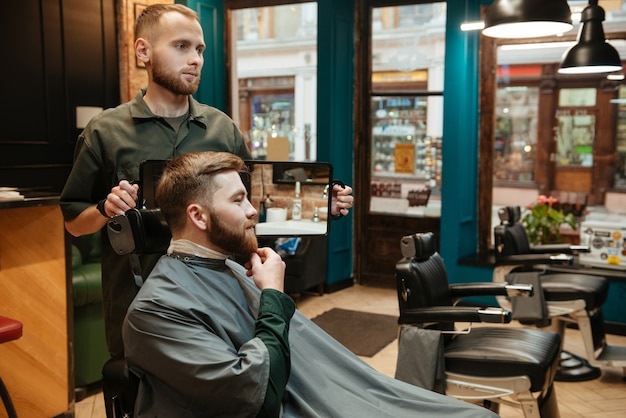 jovem bonito cortando cabelo de barba de cabeleireiro enquanto está sentado na cadeira de barbearia.