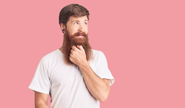 Jovem bonito de cabeça vermelha com barba longa vestindo camiseta branca casual com a mão no queixo pensando em pergunta expressão pensativa sorrindo com rosto pensativo conceito de dúvida