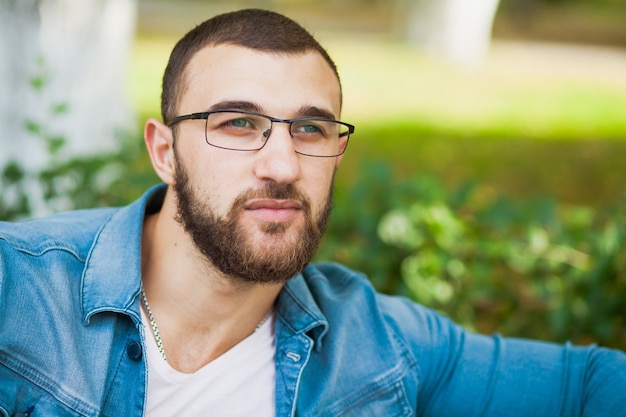 Jovem bonito de óculos com barba relaxar ao ar livre. closeup retrato masculino de um cara com jaqueta jeans, estilo jeans, aparência hipster