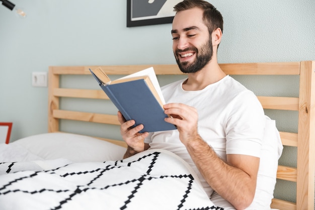 Jovem bonito deitado na cama, trabalhando em um laptop, lendo um livro